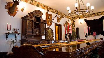A formal dining room with a long table, ornate chairs, a chandelier, and wall-mounted deer antlers.