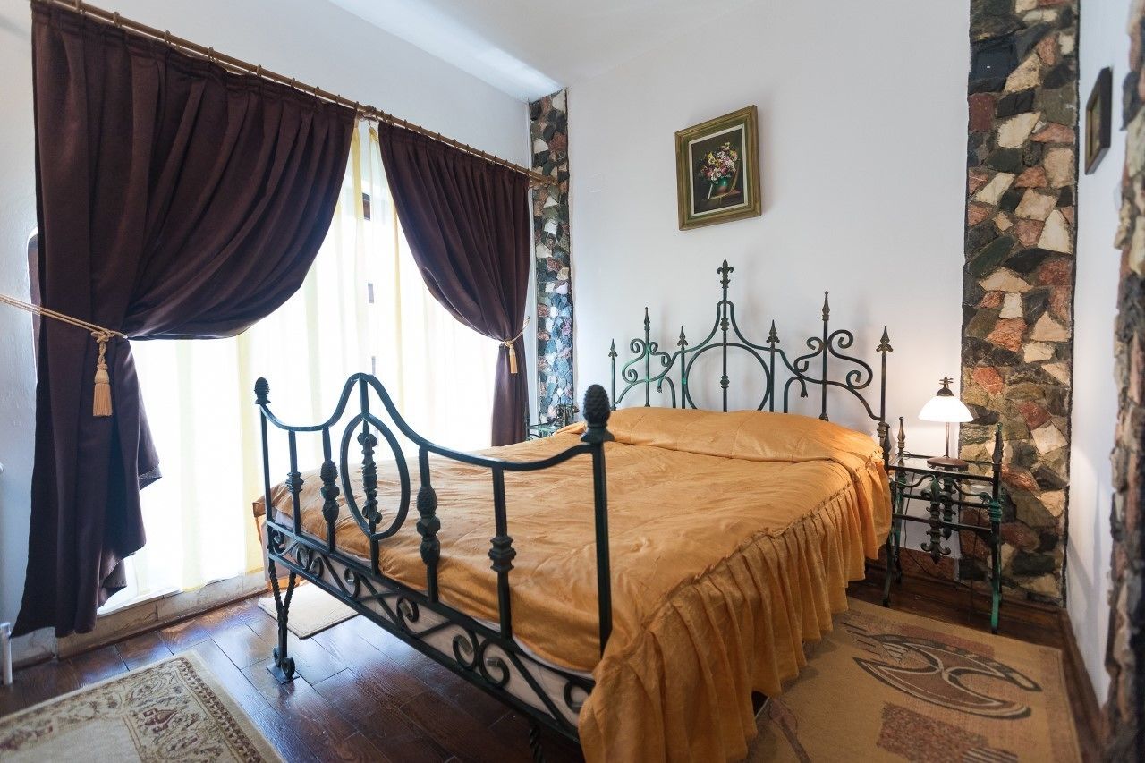 A bed with a metal frame and a yellow bedspread, a stone wall, a small table with a lamp, and a window with curtains.