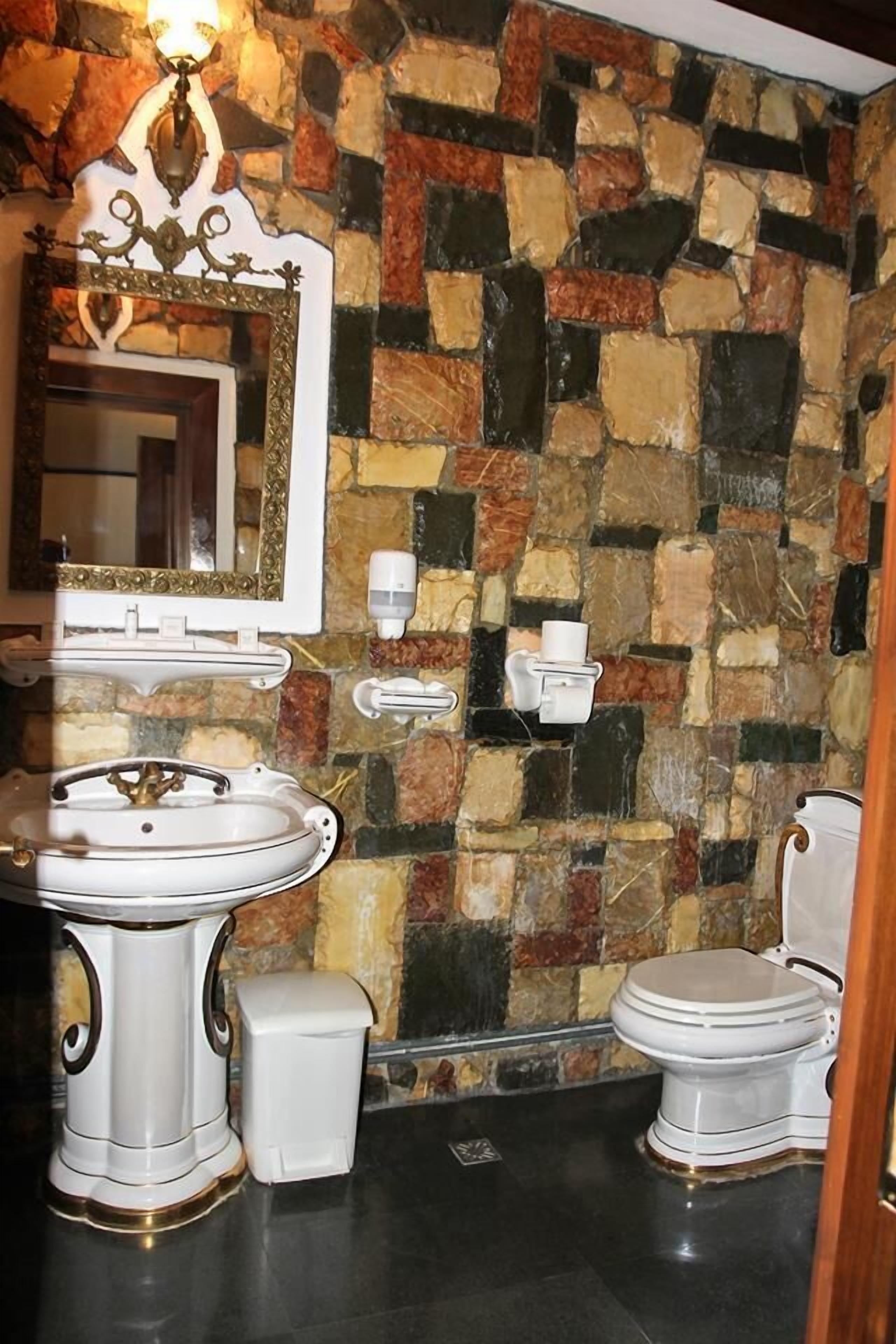 Un baño con pared de piedra, lavamanos pedestal, inodoro y espejo con marco decorativo.