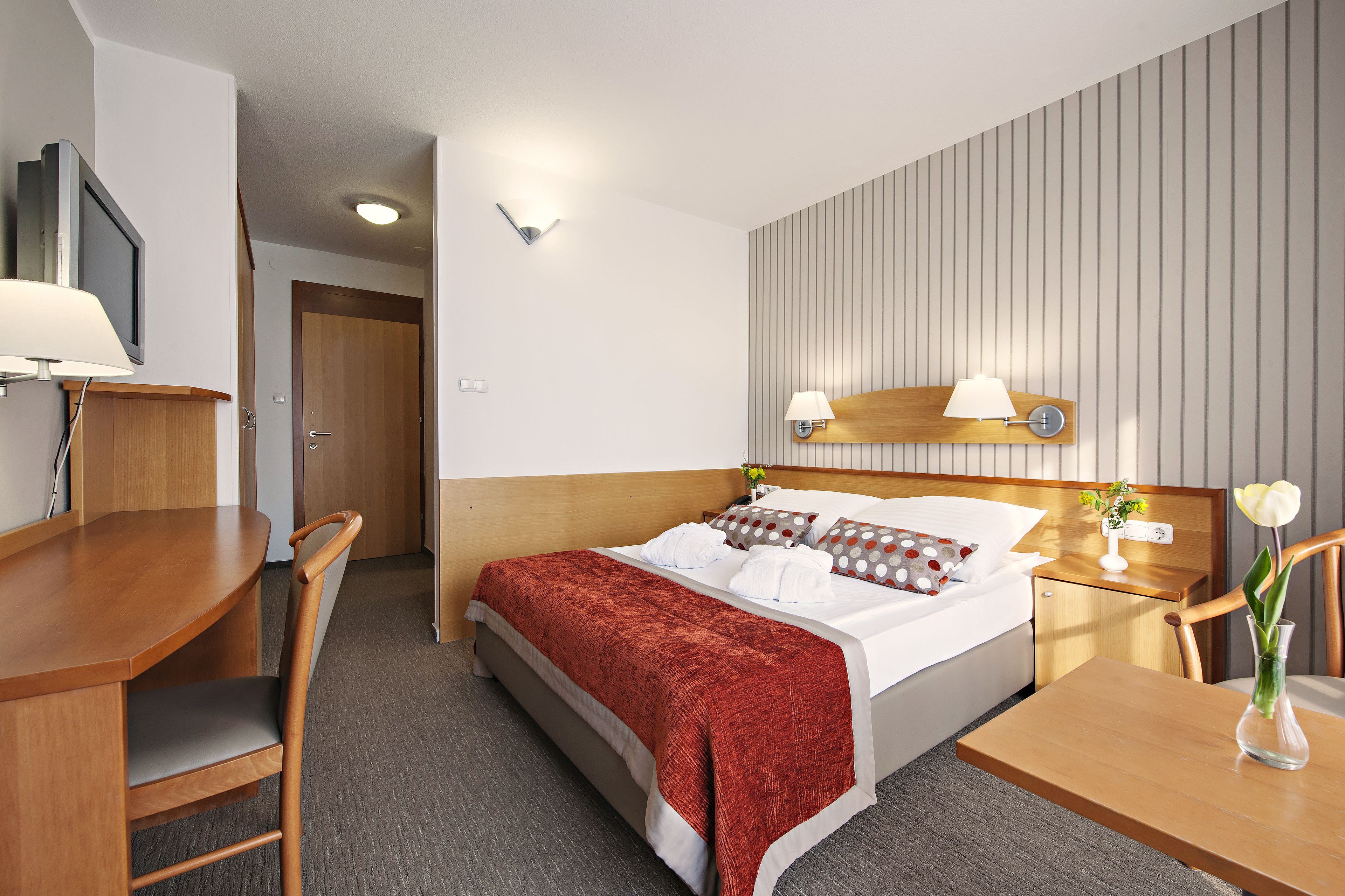 Habitación de hotel con cama, mesita de noche, lámpara, silla y escritorio con otra silla.