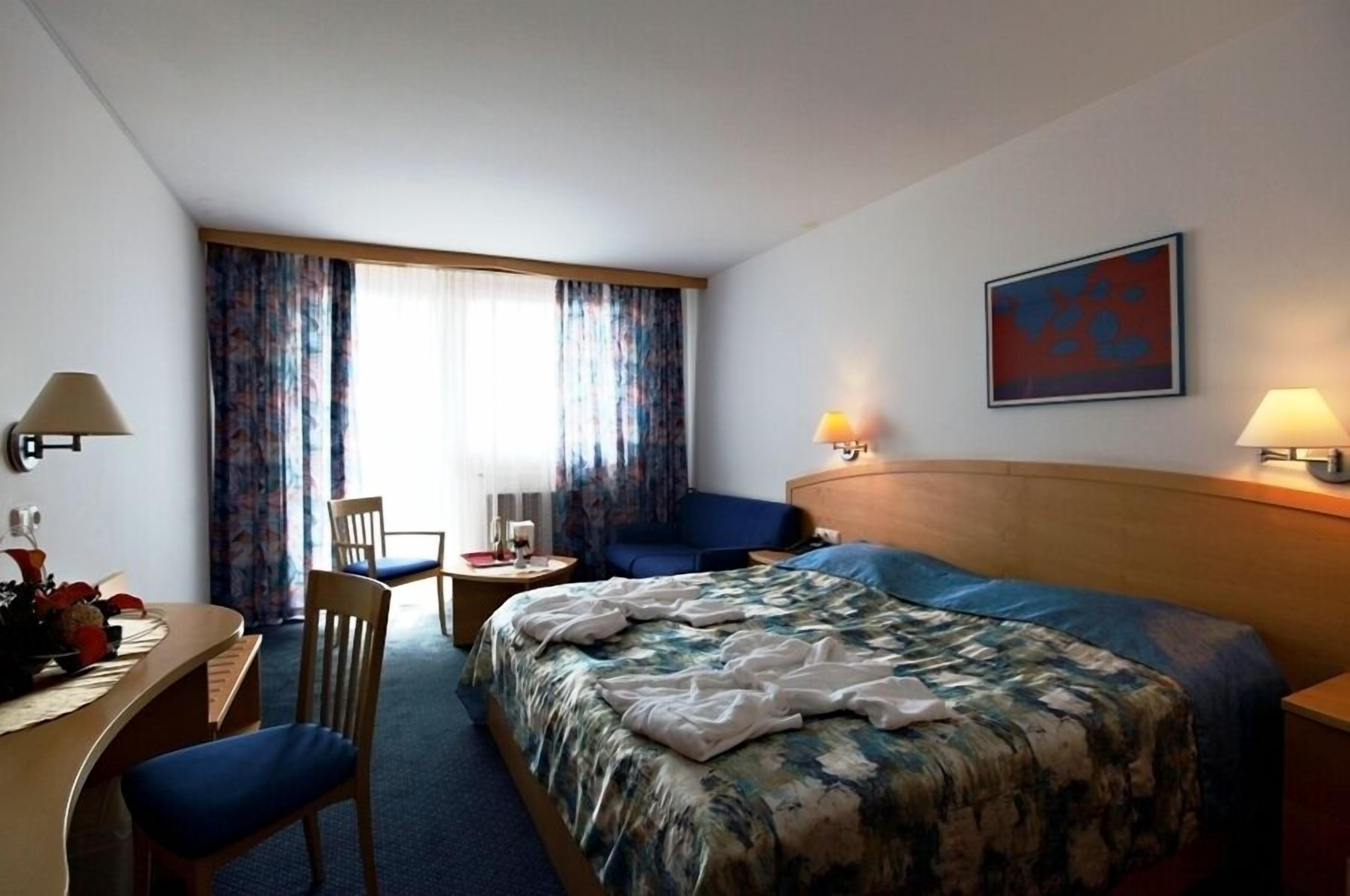 A hotel room with a bed, a desk, a chair, a small table, and a window with curtains.