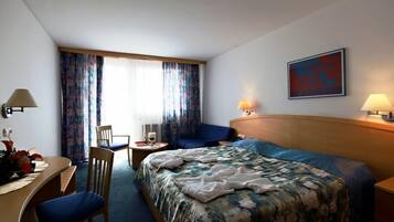 A hotel room with a bed, a desk, a chair, a small table, and a window with curtains.