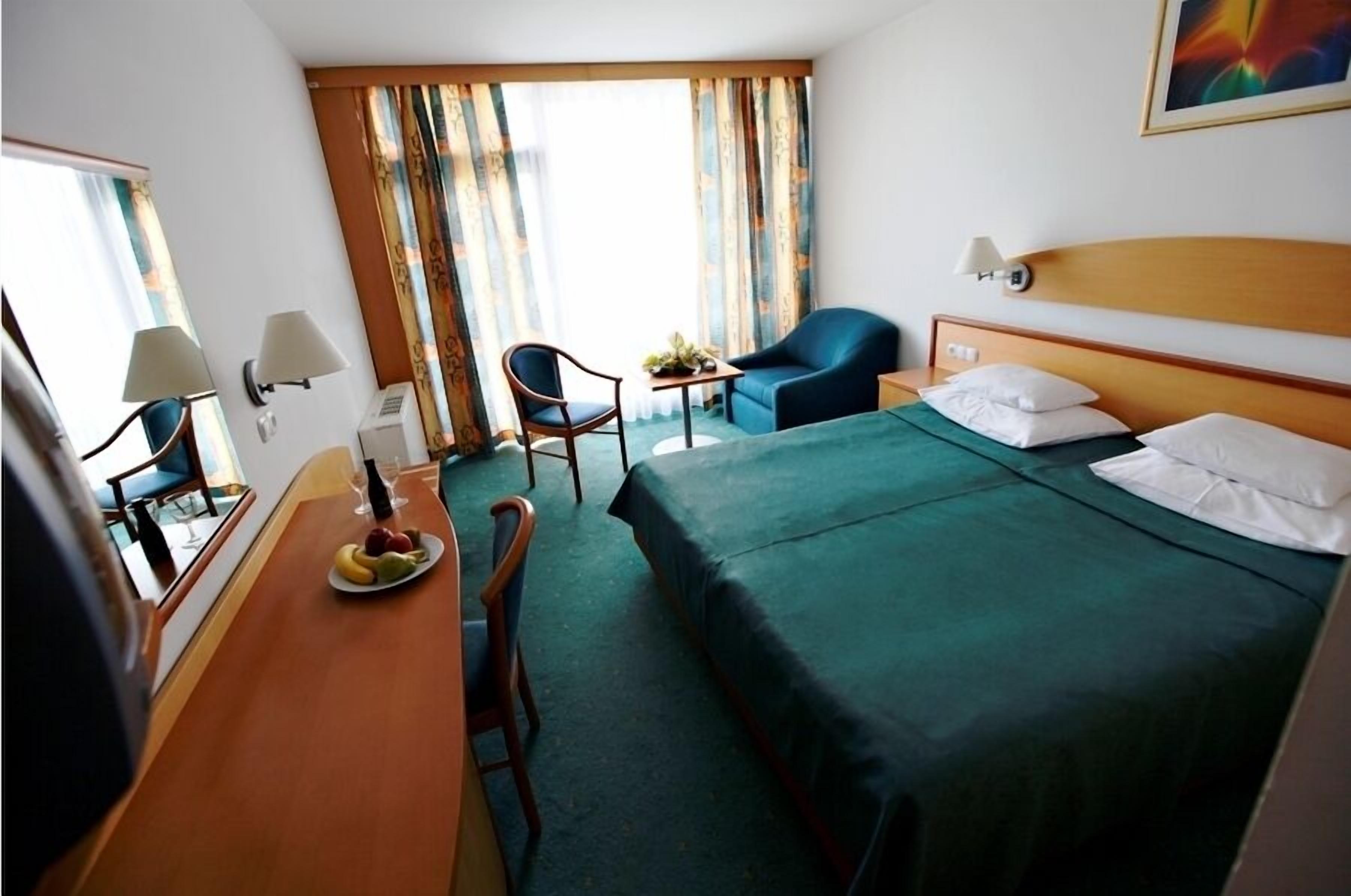 A hotel room with a bed, a desk with a chair, a small table with a chair, and a window with curtains.
