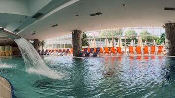 Une piscine intérieure dotée de plusieurs couloirs, d’une cascade et de chaises longues orange disposées tout autour.