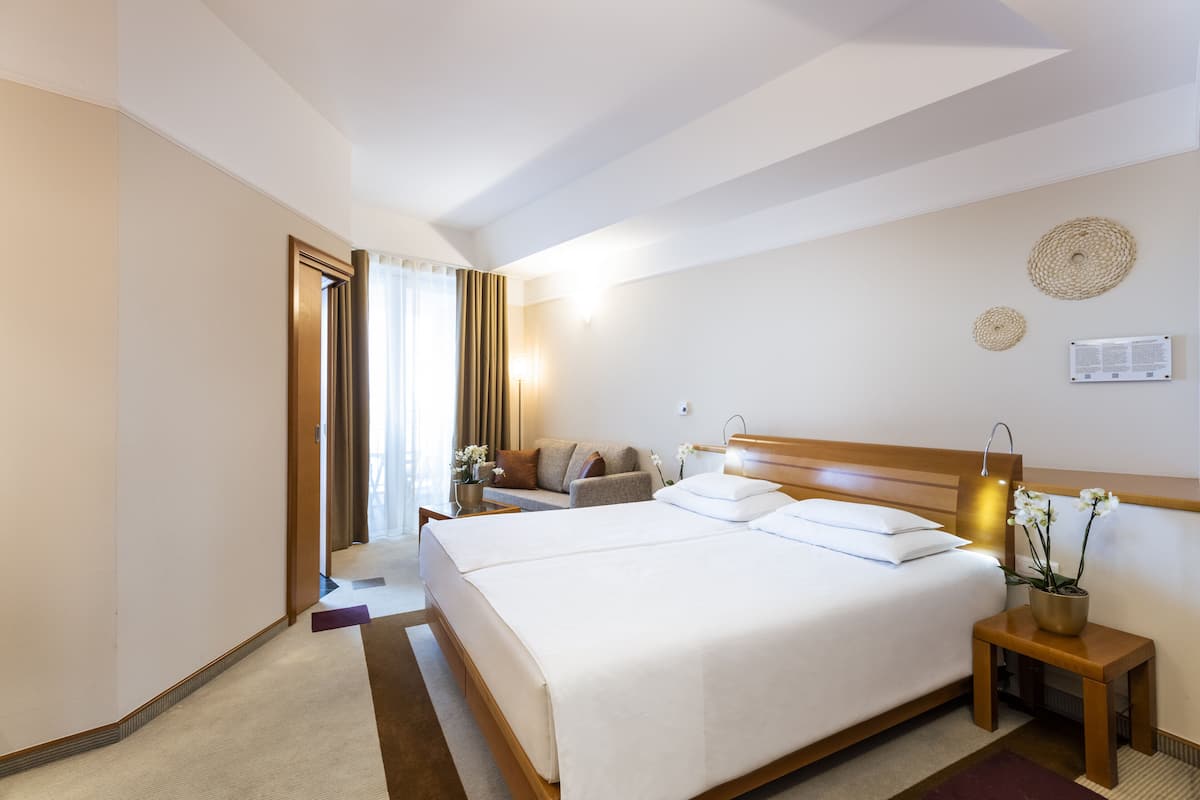 Une chambre d’hôtel avec un grand lit, une table de chevet avec une lampe, une petite table avec des fleurs et une vue sur le couloir.