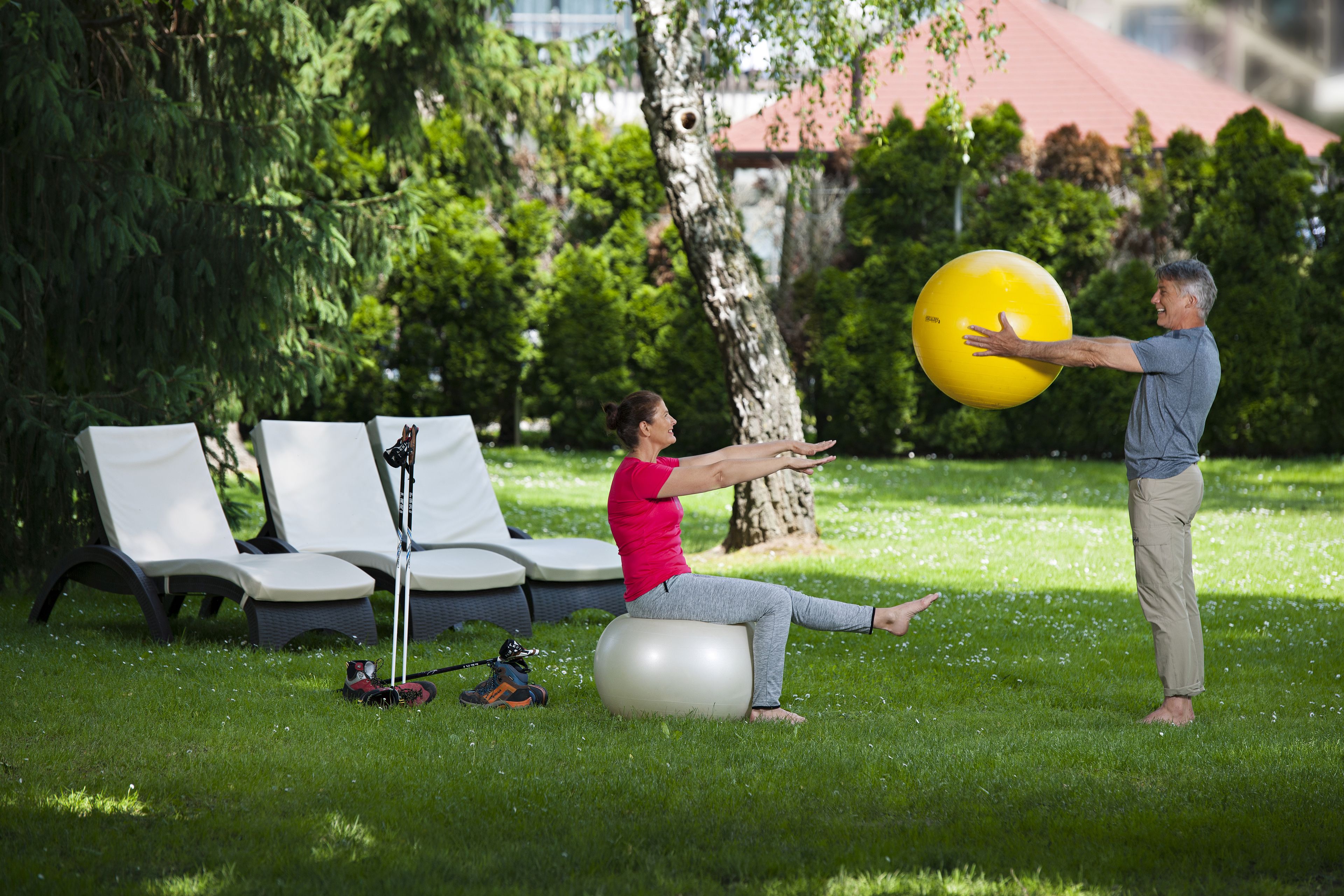 A person sitting on an exercise ball in a grassy area with lounge chairs and a tree in the background.