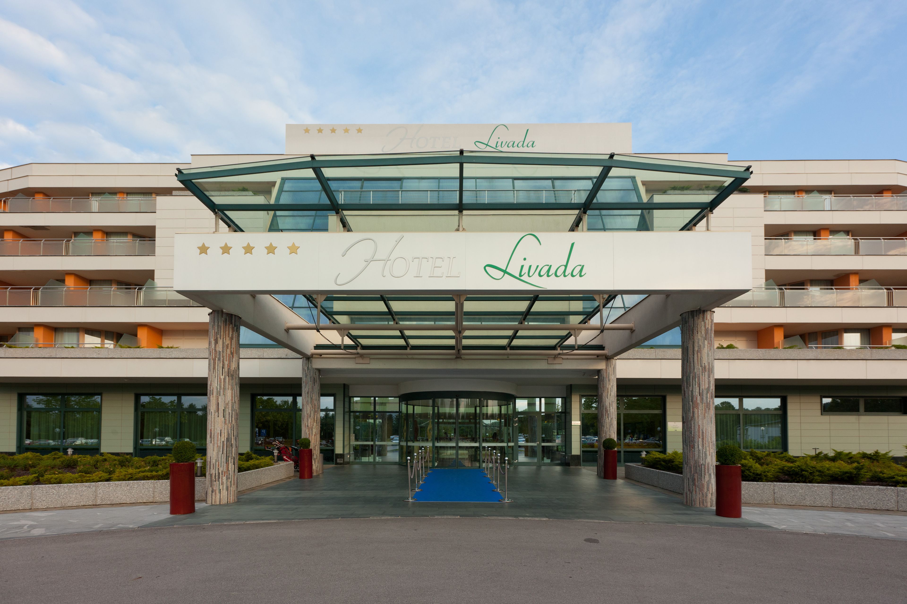 A modern hotel entrance with a glass canopy, a blue carpet, and a visible name sign.