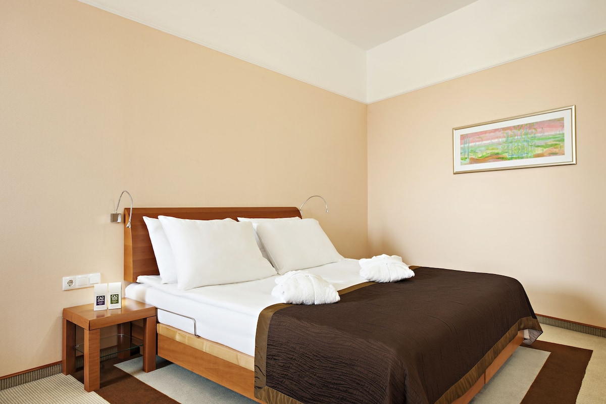 Un lit bien fait, avec des draps blancs et une couverture marron, une table de chevet en bois avec une lampe, et un tableau encadré accroché au mur.