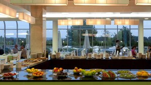 A buffet setup with various fruits and beverages, large windows offering a view of the outdoors, and a spacious dining area with tables and chairs. - Hotel Livada Prestige - Sava Hotels & Resorts (Moravske Toplice)