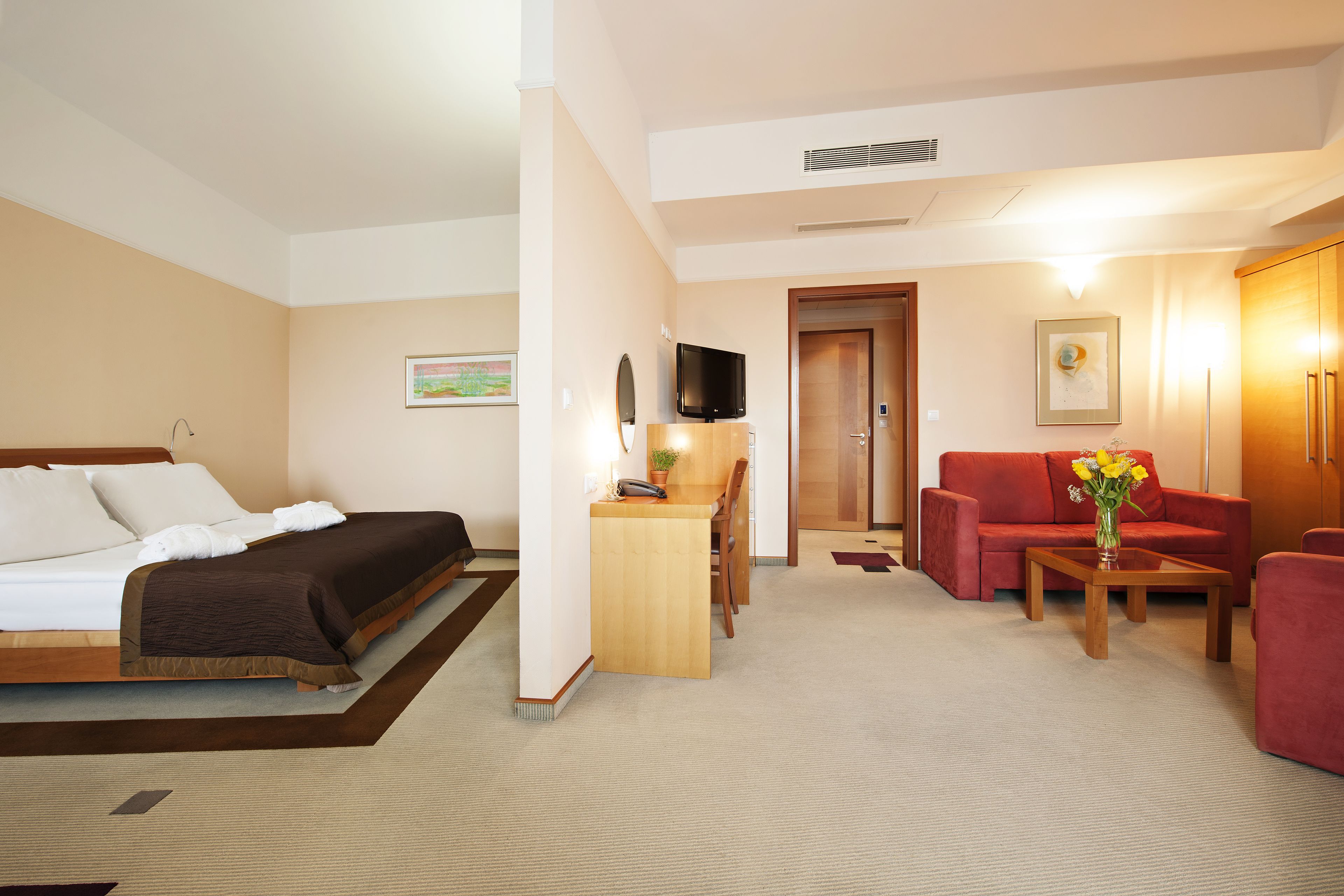A hotel room with a bed, a desk, a red sofa, and a small table with flowers.