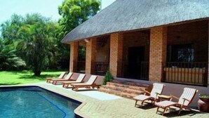 A poolside area with lounge chairs and a building with a thatched roof. - Dreamfields Guesthouse (Mbombela)