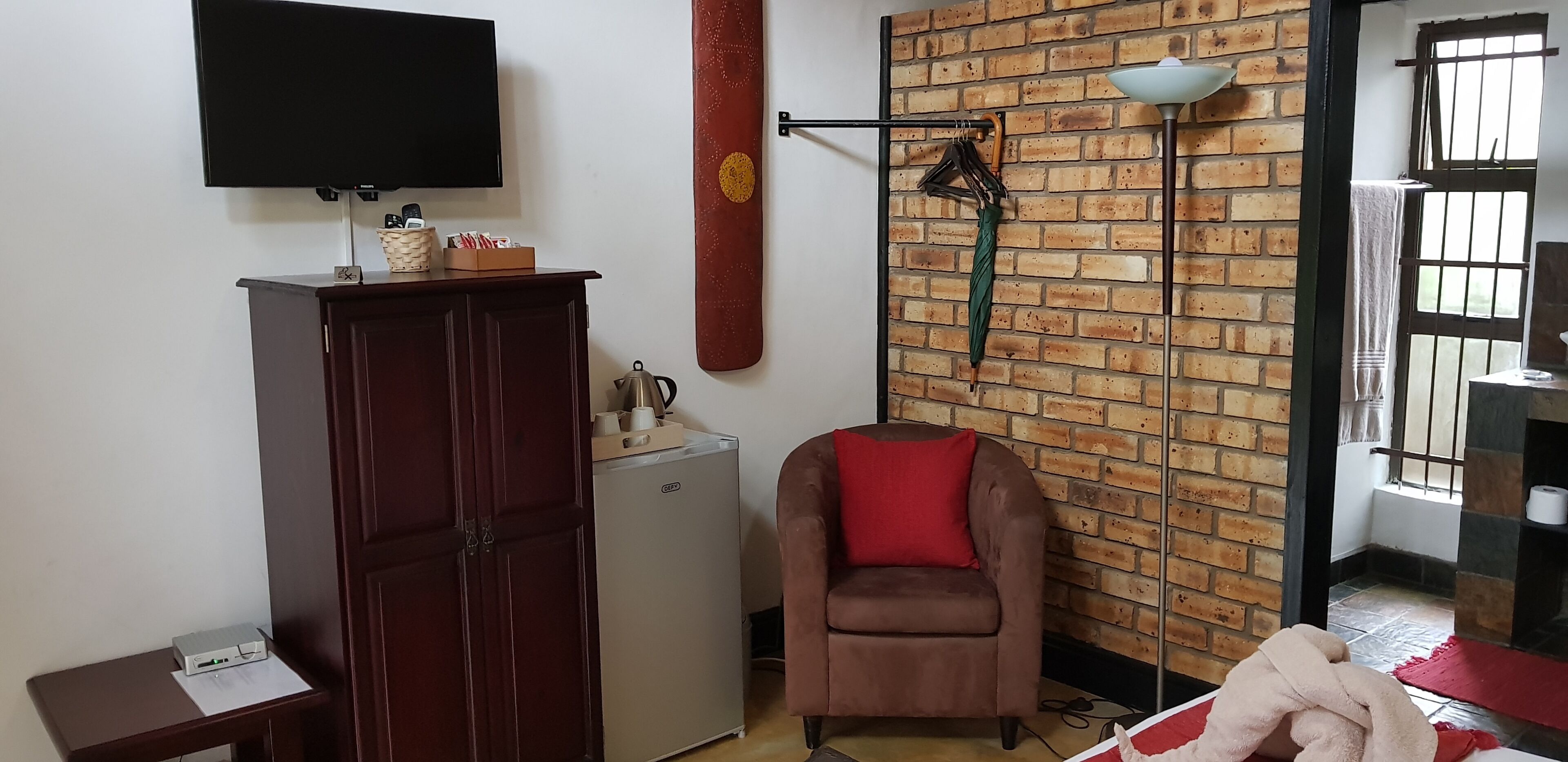 A room with a brick wall, a flat-screen TV mounted above a wooden cabinet, a small refrigerator, a brown armchair with a red cushion, and a window with bars.