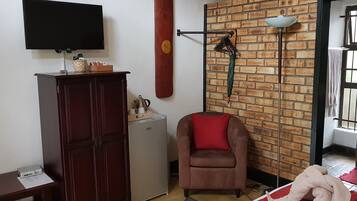 A room with a brick wall, a flat-screen TV mounted above a wooden cabinet, a small refrigerator, a brown armchair with a red cushion, and a window with bars.