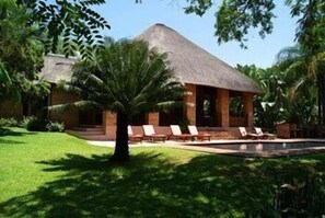 A house with a thatched roof, a swimming pool, and lounge chairs. - Dreamfields Guesthouse (Mbombela)