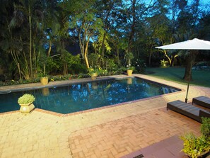 A rectangular swimming pool with a tiled edge, surrounded by a brick patio. There are potted plants and a white umbrella on the patio. - Dreamfields Guesthouse (Mbombela)