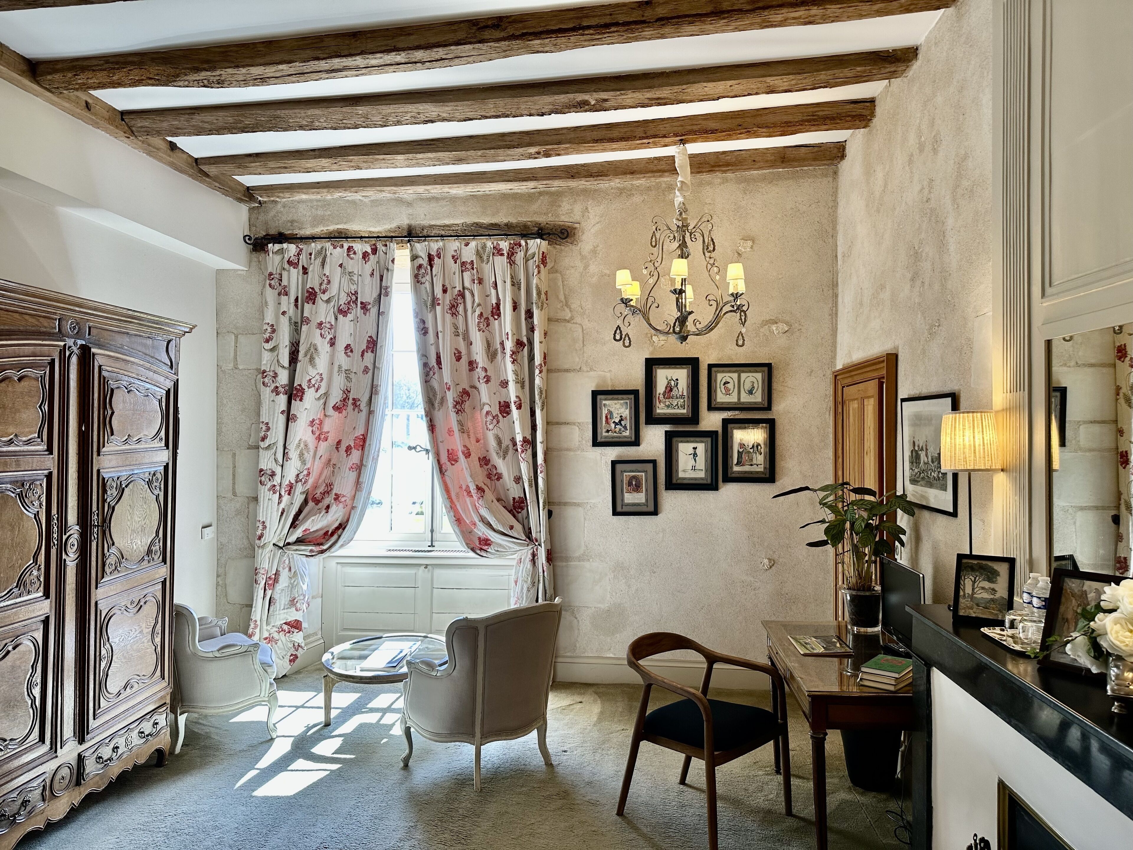 A room with a wooden ceiling, a chandelier, a window with floral curtains, a wooden cabinet, a chair, a table, and framed pictures on the wall.