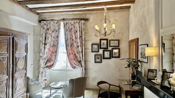 A room with a wooden ceiling, a chandelier, a window with floral curtains, a wooden cabinet, a chair, a table, and framed pictures on the wall.