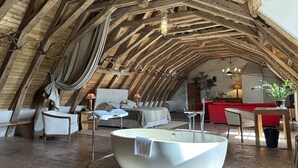 An attic bedroom with a large bathtub, wooden beams, a red sofa, and a chandelier. - Château des Arpentis (Saint-Regle)
