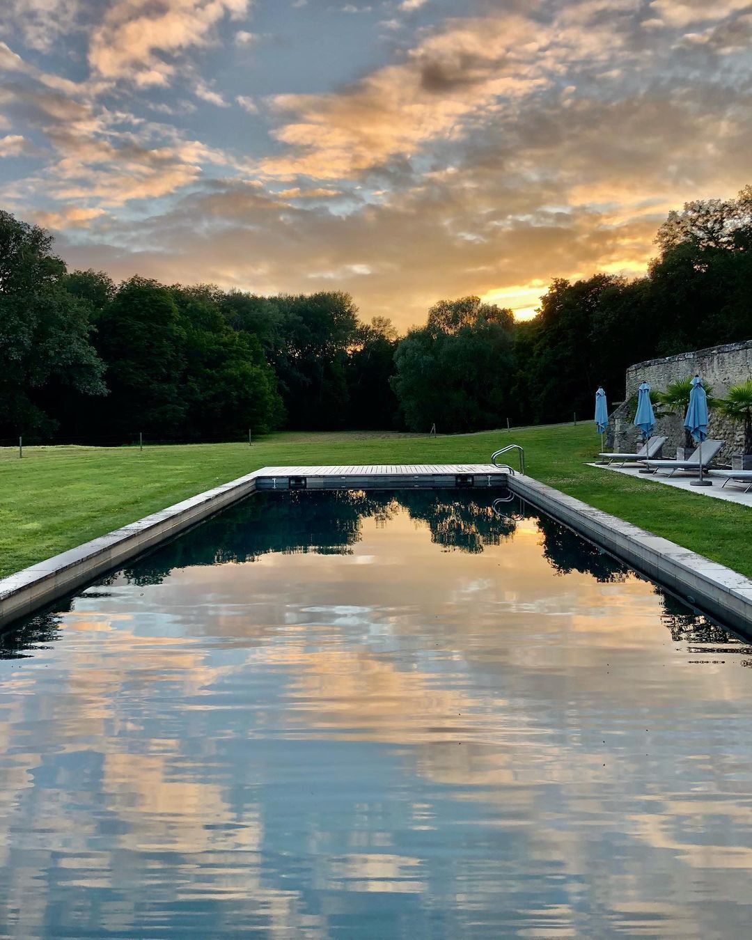 A rectangular swimming pool with a wooden deck in the center, surrounded by a grassy area and trees.