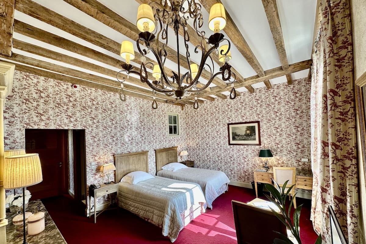 A bedroom with a wooden ceiling, floral wallpaper, a chandelier, a bed with two pillows, a nightstand, a desk, a chair, and a window.