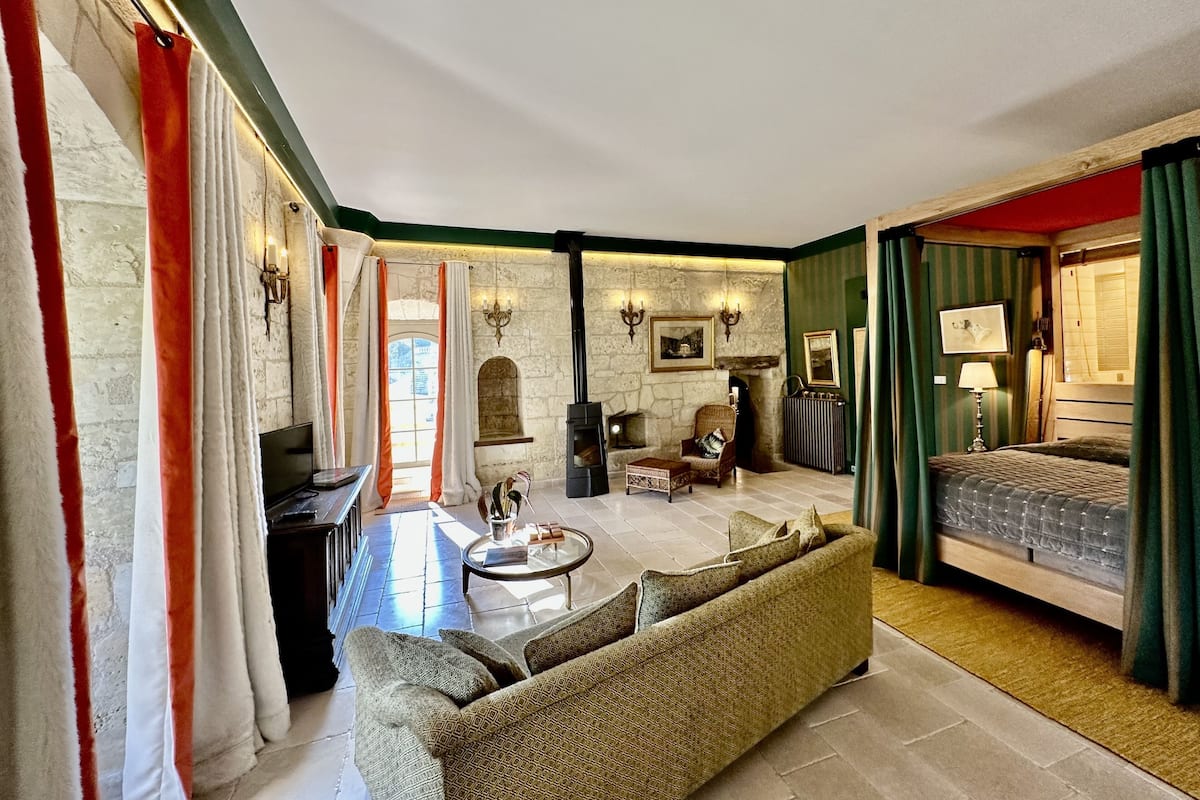 A cozy living room with a sofa, armchair, and coffee table, a fireplace, and a bed with green curtains.