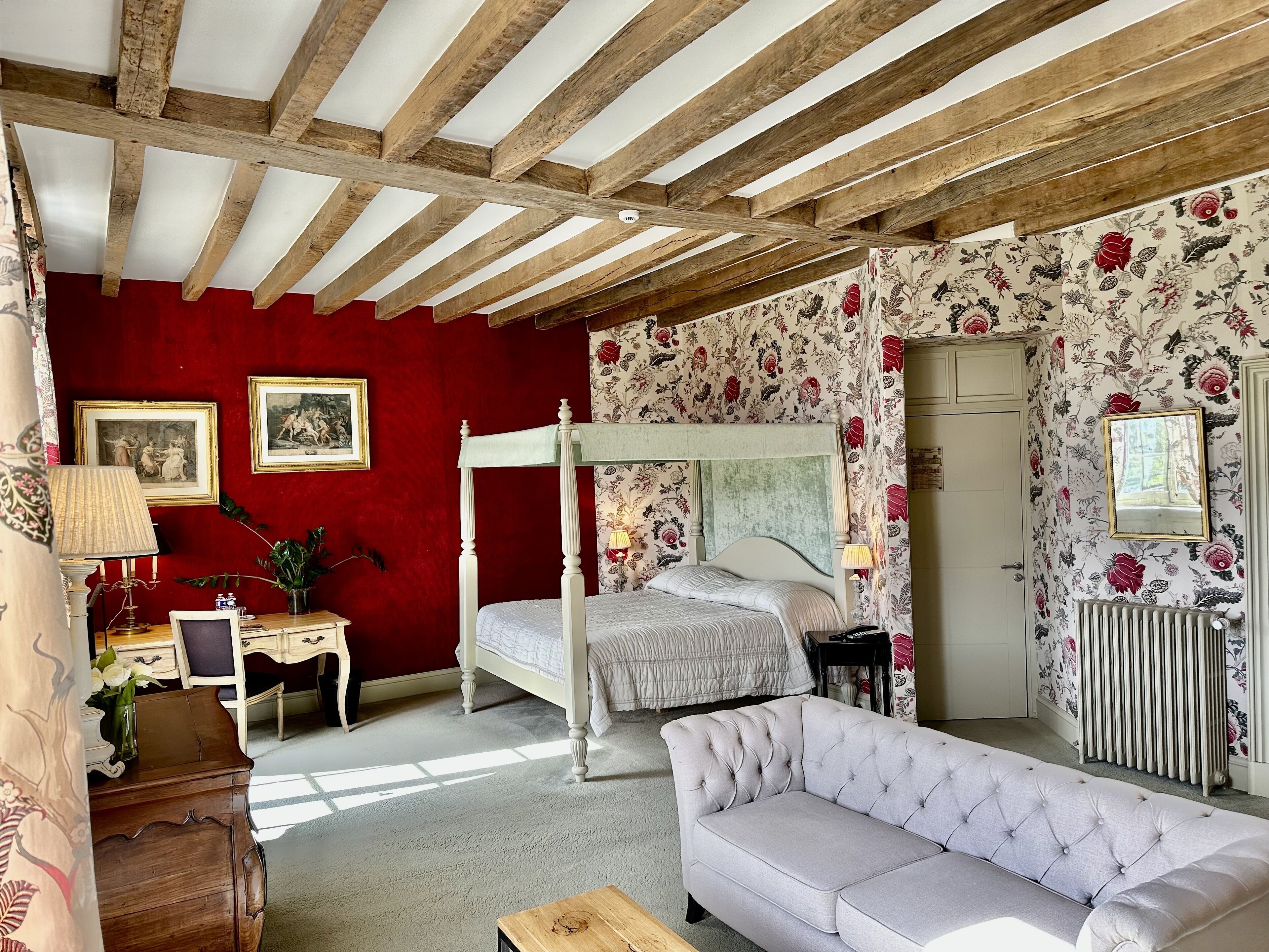 A bedroom with a four-poster bed, a sofa, a desk with a lamp, and floral wallpaper.