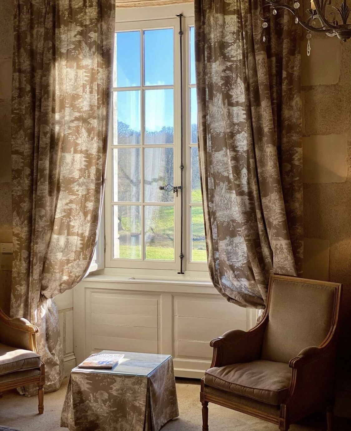 A room with a large window, two chairs, a small table, and a chandelier.