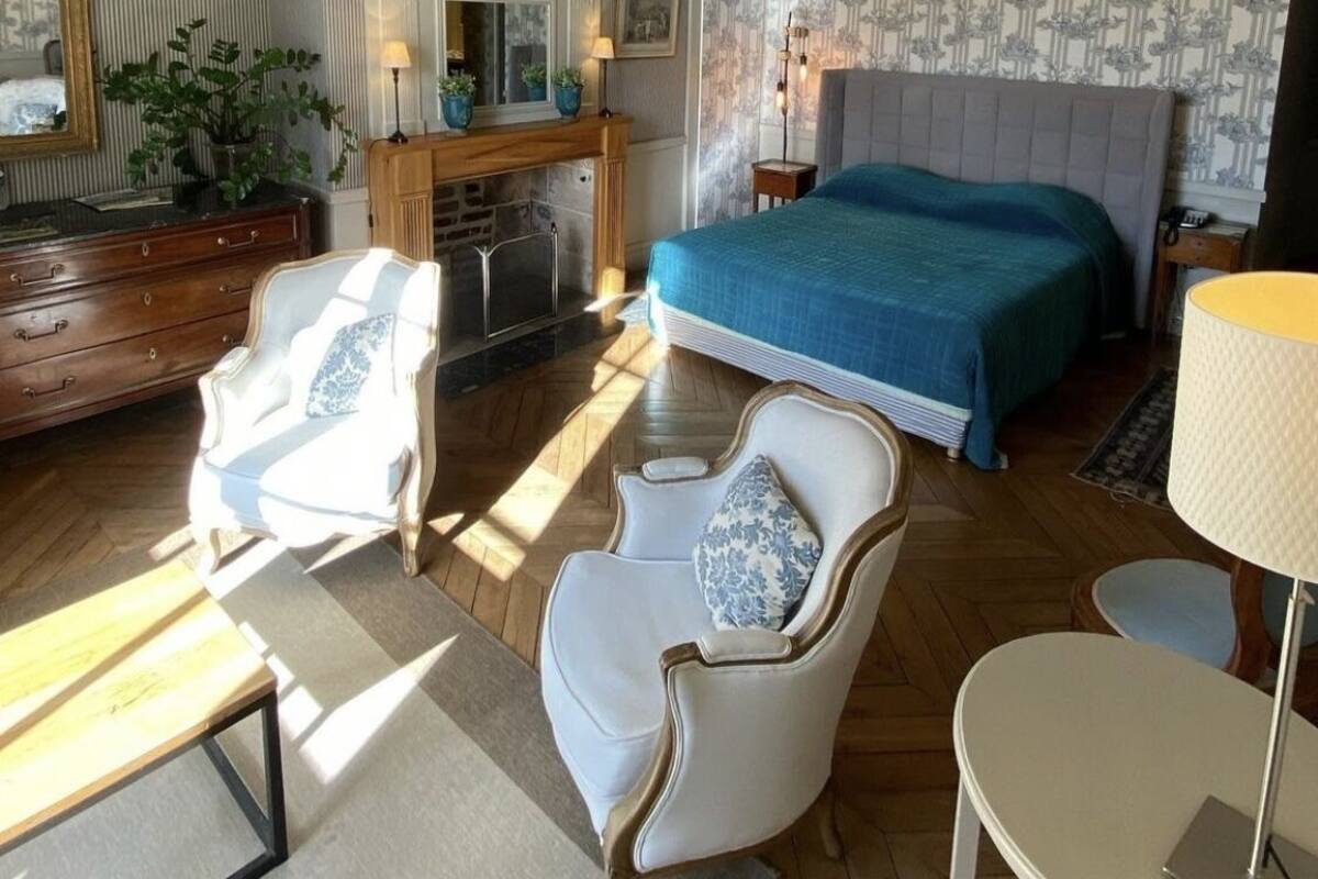 A spacious bedroom with a large bed, a fireplace, and a chandelier. There are two white armchairs and a small table with a lamp.