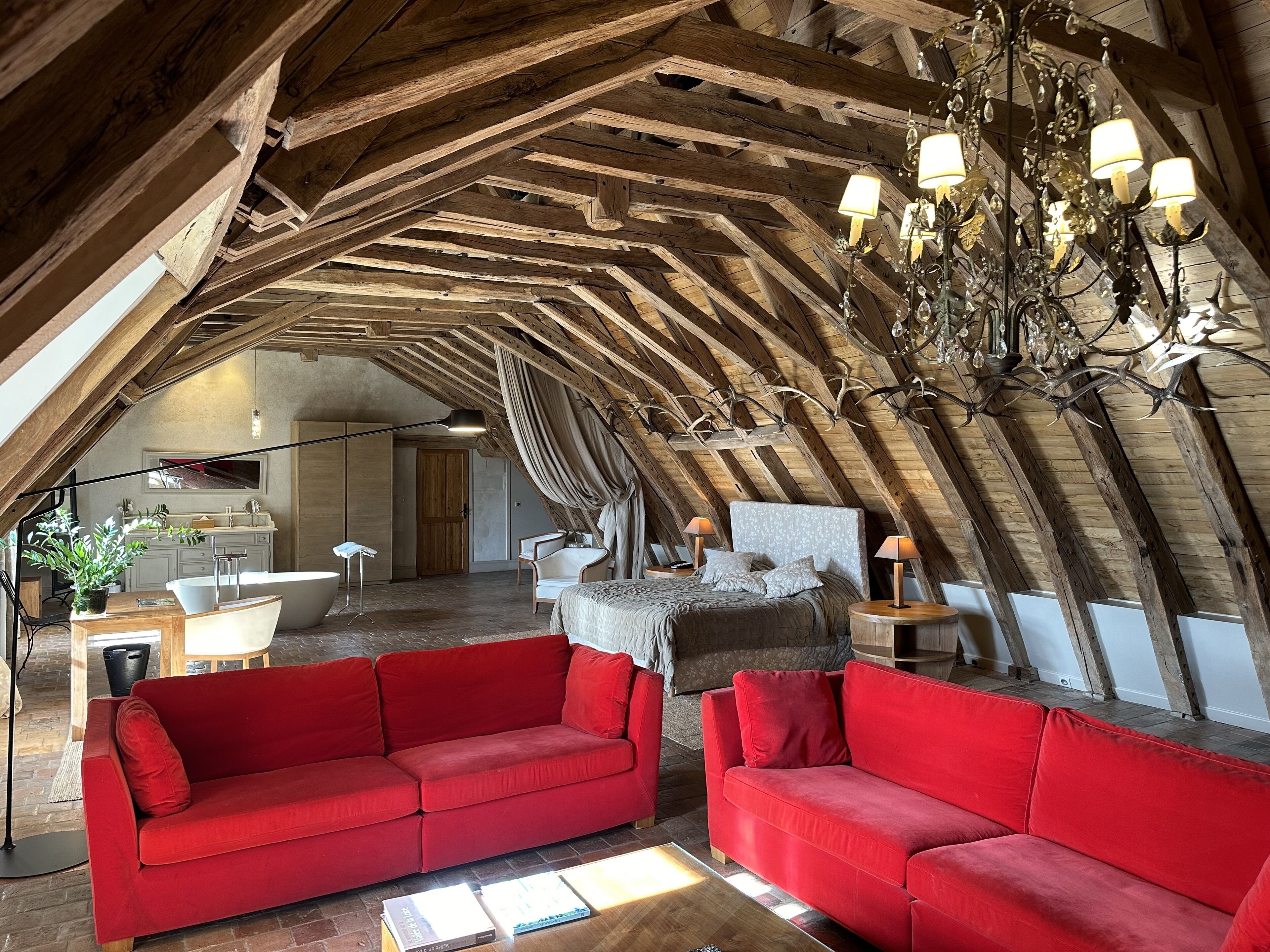 Un acogedor dormitorio en el ático con techo inclinado, vigas de madera, un candelabro, un sofá rojo y una cama con cabecera blanca.