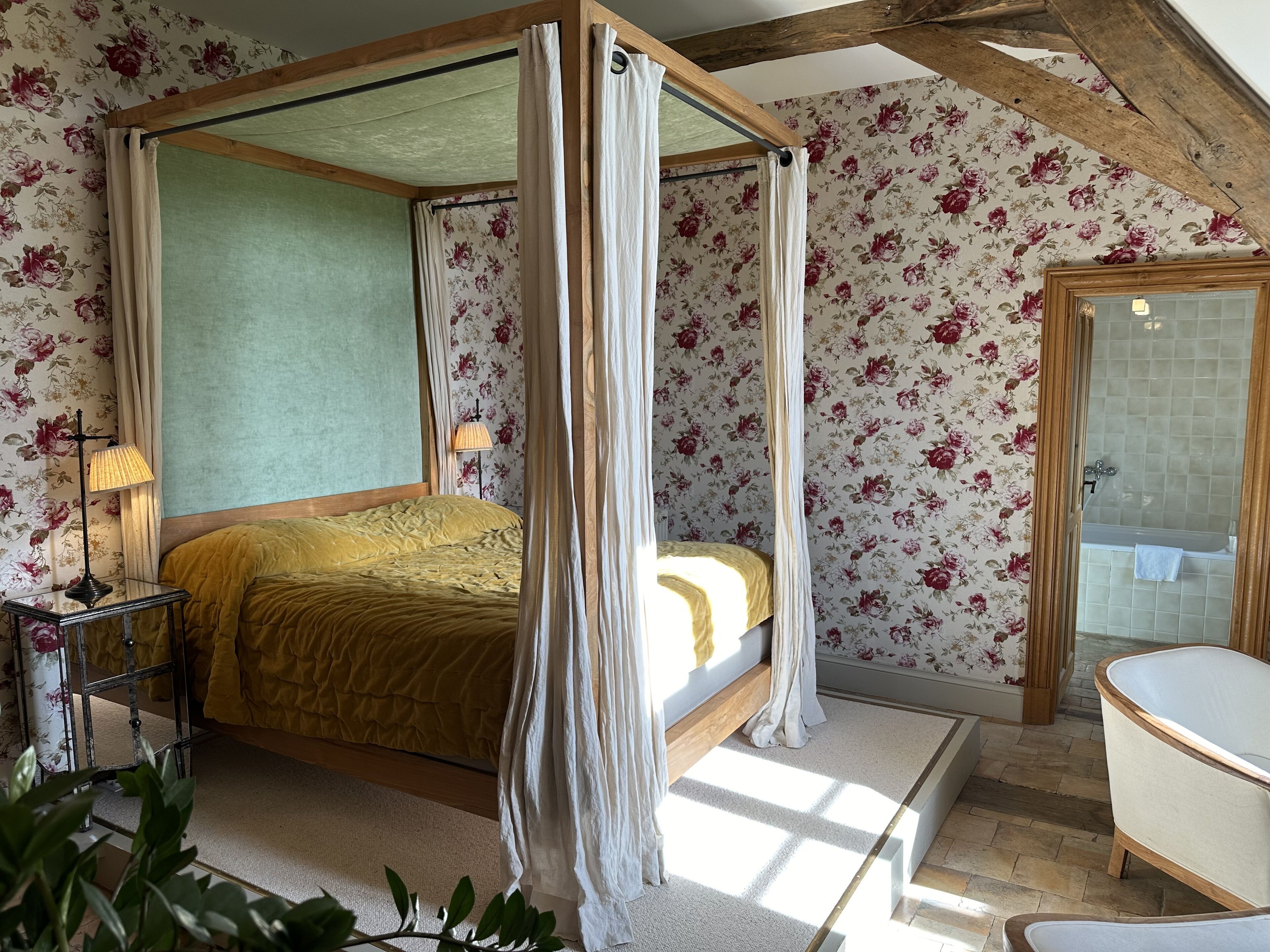 Una cama con dosel sobre cuatro pilares, con un marco de madera, una mesita de noche con lámpara y un estampado floral en el papel tapiz.