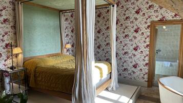 Una cama con dosel sobre cuatro pilares, con un marco de madera, una mesita de noche con lámpara y un estampado floral en el papel tapiz.