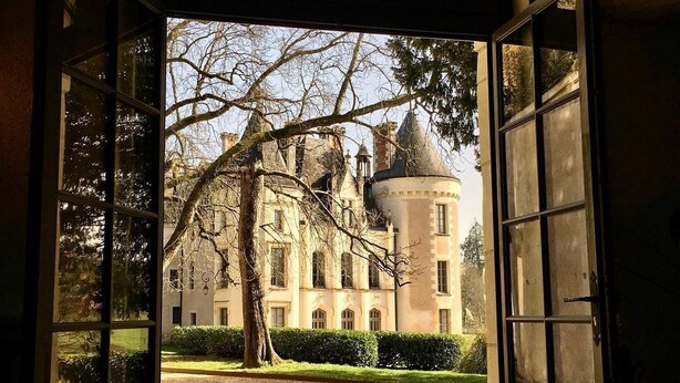 A view through an open arched doorway leading to a courtyard with a large tree and a building with a dome. - Château des Arpentis (Saint-Regle)
