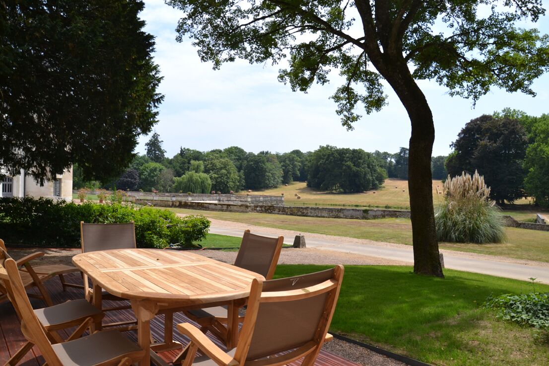 An outdoor patio with a wooden table and chairs, surrounded by greenery and a view of a park.