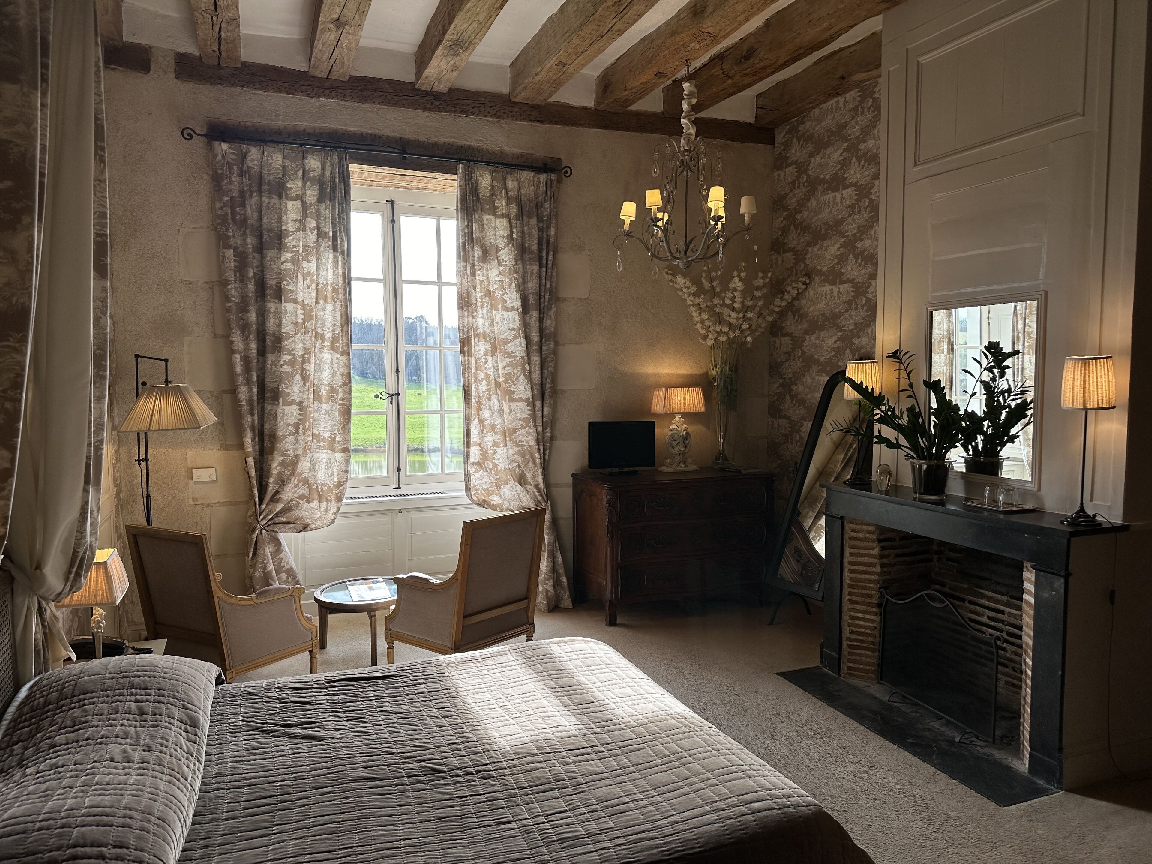Ein Schlafzimmer mit einem großen Bett, einem kleinen Tisch, einem Stuhl, einem Kamin und Blick ins Freie.