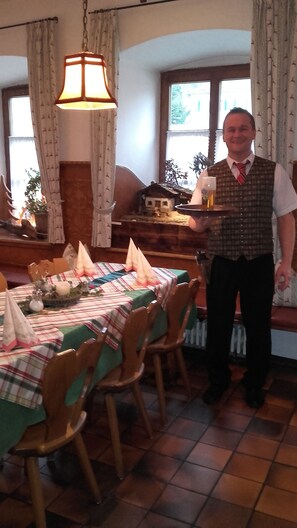 A dining area with a table set for four, a hanging light fixture, and a window with curtains. - Gasthof zum Rassen (Garmisch-Partenkirchen)