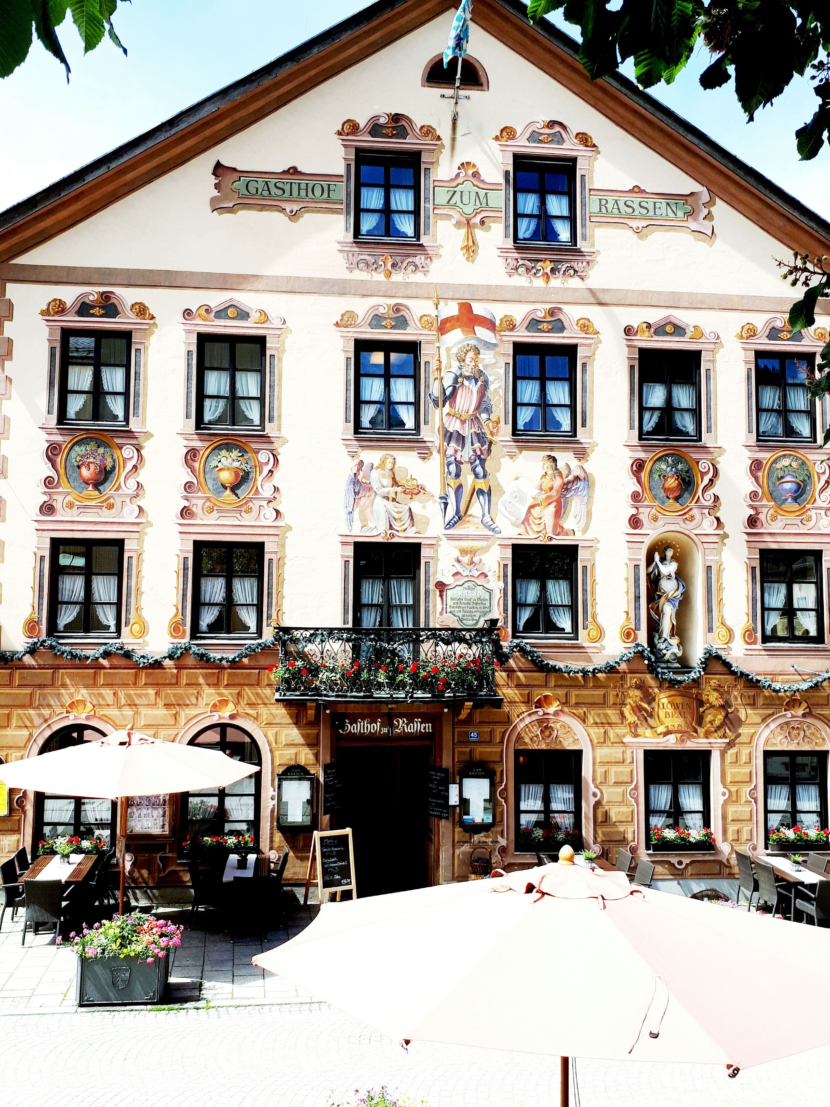 Ein Gebäude mit aufwendigen architektonischen Details, darunter ein Balkon mit Blumen und ein Schild mit der Aufschrift 'Gasthof Zum Rassen'.