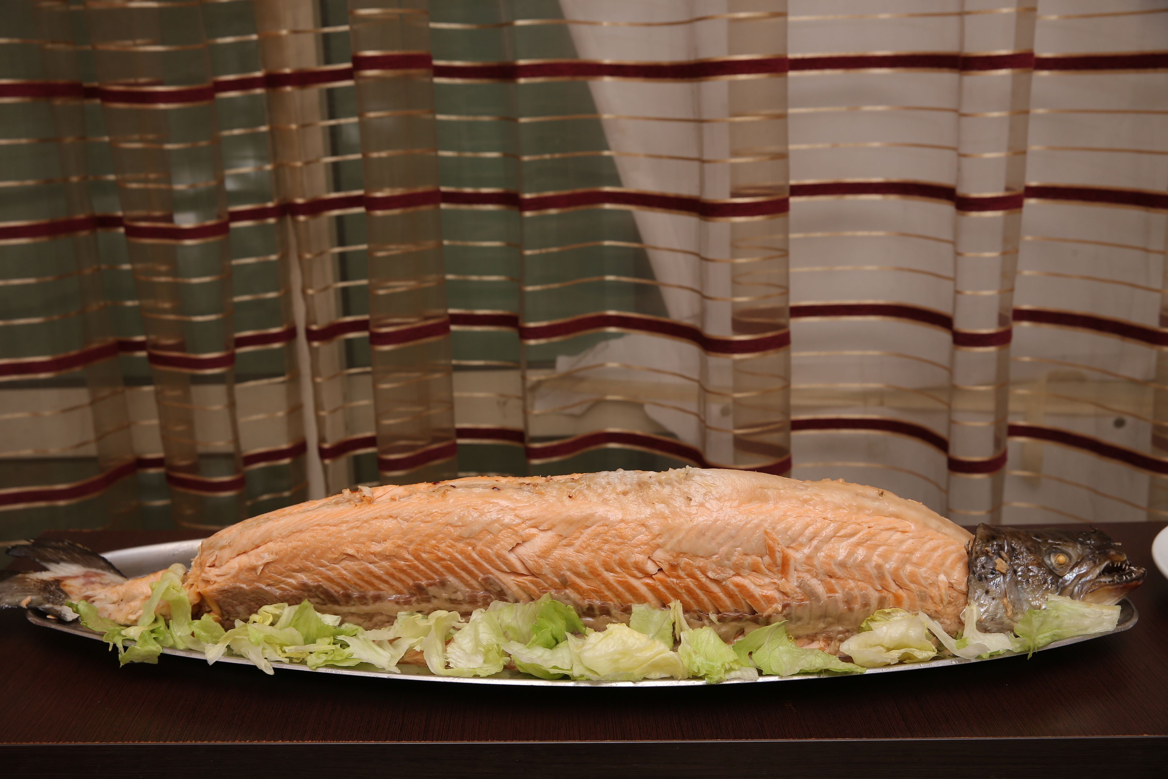 A whole cooked fish on a plate with lettuce, set on a table with a striped curtain in the background.
