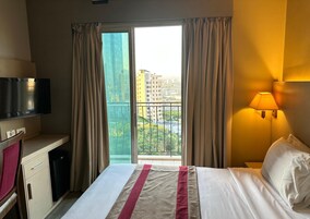Habitación de hotel con cama, televisión, escritorio, lámpara y vistas a edificios por la ventana.