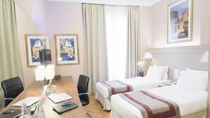 A hotel room with a wooden desk, two beds, a large window with curtains, and framed artwork on the walls. - Padova Hotel (Sin El Fil)