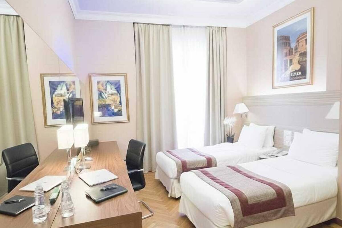 Habitación de hotel con un escritorio de madera, dos camas, un ventanal con cortinas y obras de arte enmarcadas en las paredes.