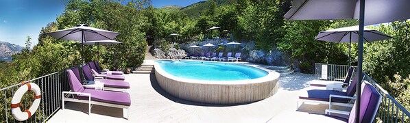 A circular hot tub surrounded by lounge chairs and umbrellas, with a pool and greenery in the background.