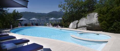 Una piscina con sillas de descanso y sombrillas, rodeada de árboles y montañas al fondo.