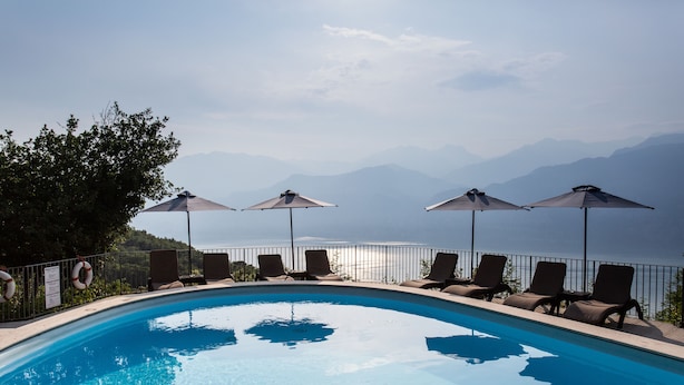 A swimming pool with lounge chairs and umbrellas, surrounded by a fence and overlooking a body of water with mountains in the background. - Hotel Querceto by Double Hospitality (Malcesine)