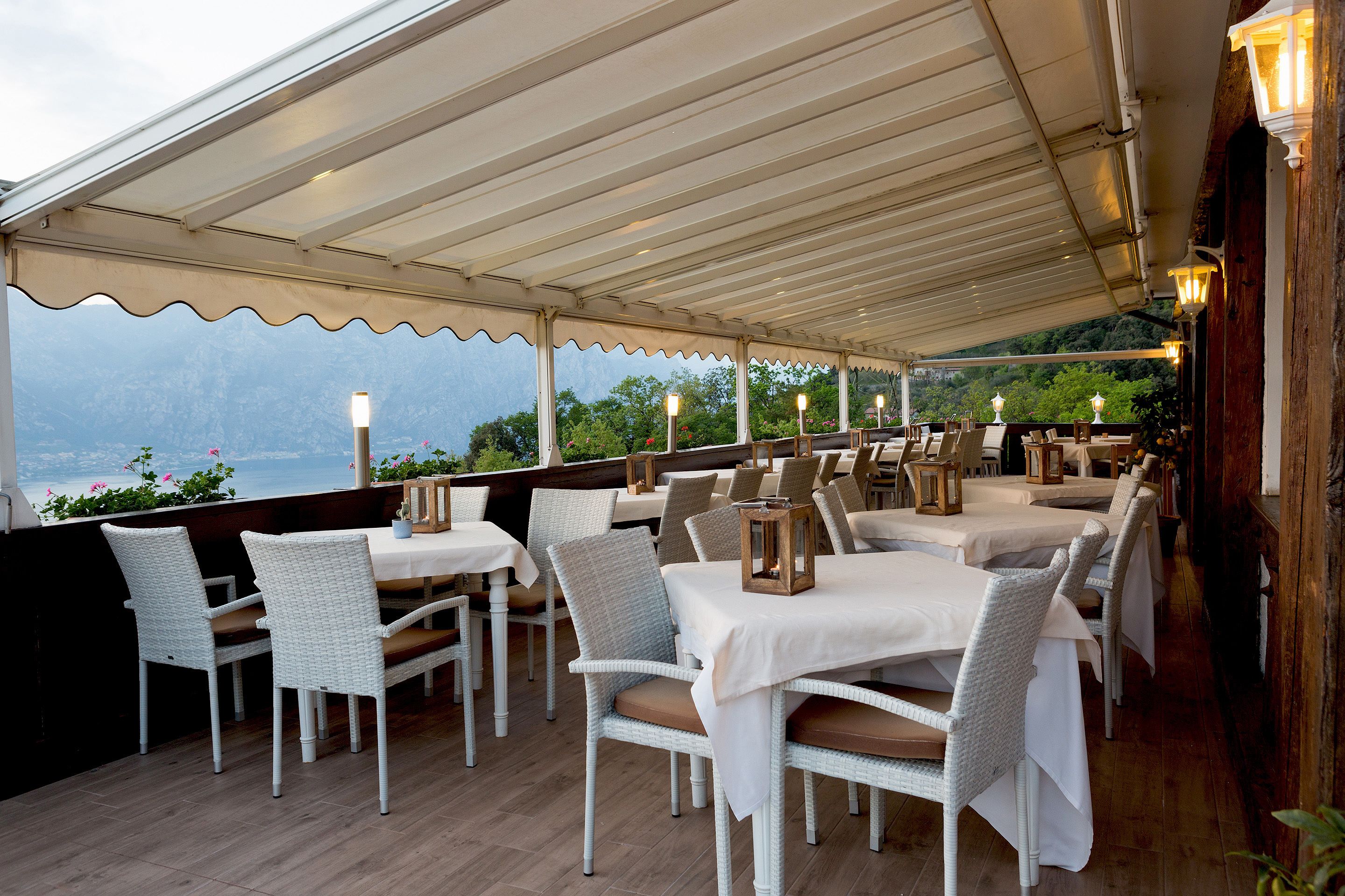 Un área de comedor al aire libre con mobiliario de mimbre blanco y mesas, cubierta por un dosel blanco, con vistas a un paisaje pintoresco.