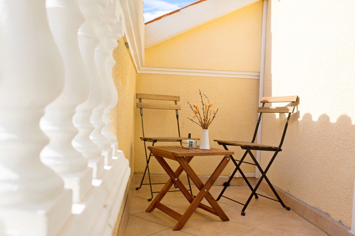 A small outdoor seating area with a wooden table and two chairs, a white vase with branches, and a yellow wall.