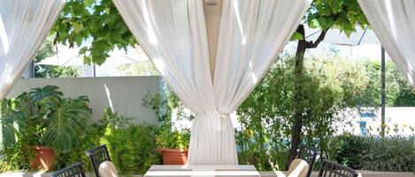 An outdoor patio with a table and chairs, surrounded by white curtains and potted plants.