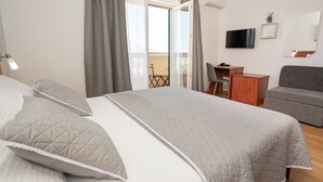 Une chambre moderne avec un grand lit, une literie grise, un bureau, un canapé gris, une télévision et un balcon avec une guitare.