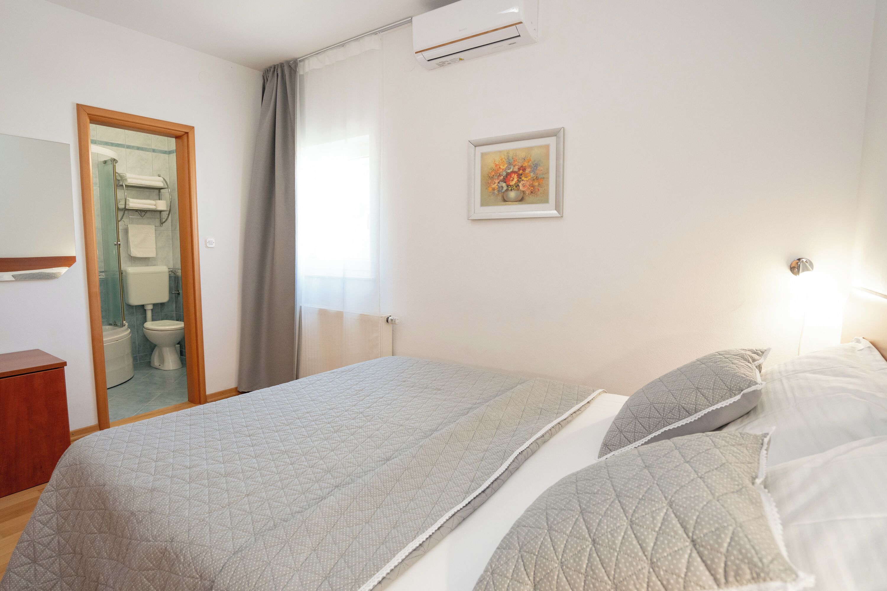 A bedroom with a bed, a wooden dresser, a bathroom visible through a glass door, and a framed picture on the wall.