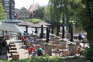 An outdoor café with tables and chairs, overlooking a body of water, with buildings in the background. - Het Rechthuis aan den Amstel (Uithoorn)