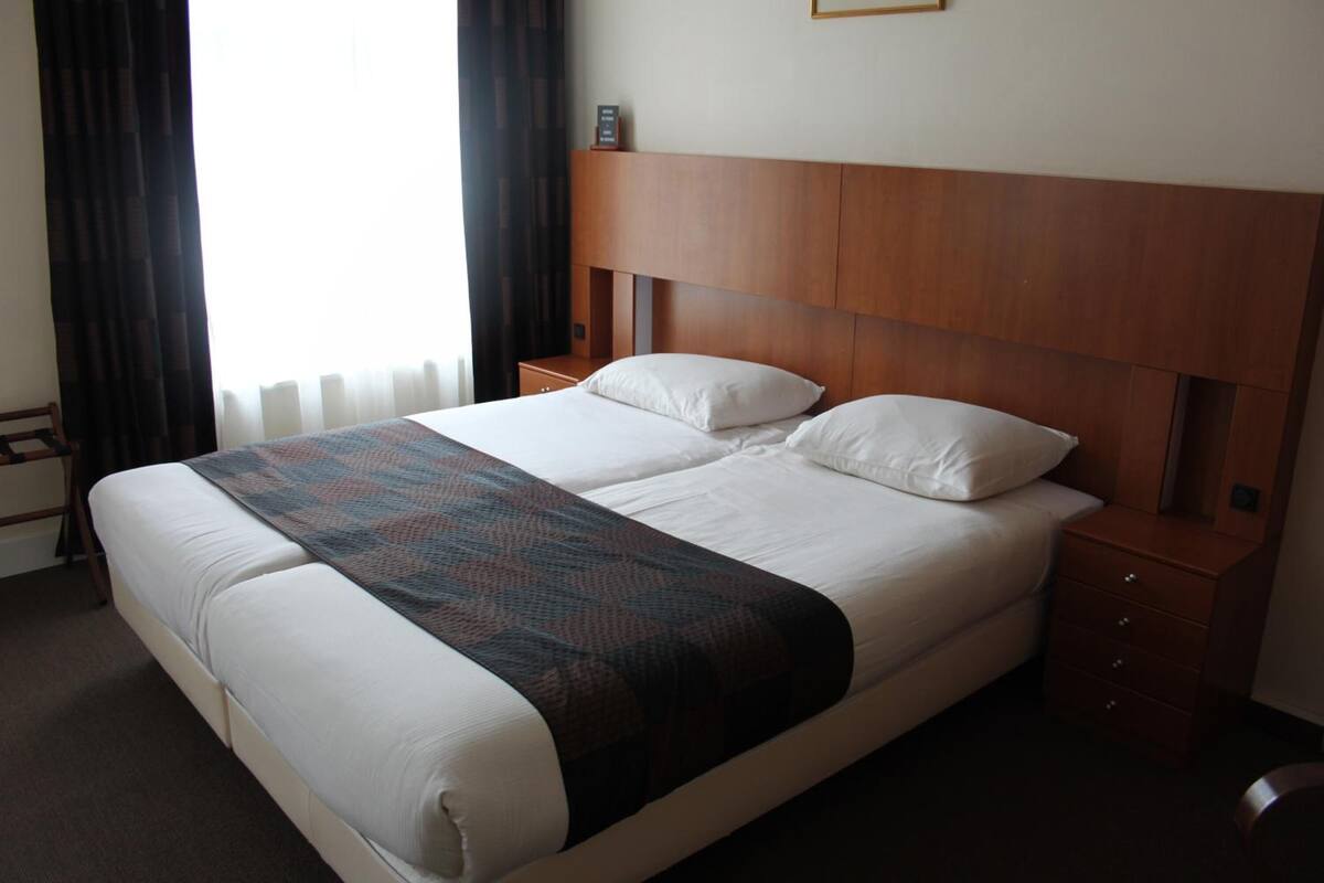 A hotel room with a large bed, a wooden headboard, a bedside table, and a window with curtains.